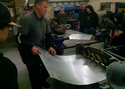 Instructor feeds sheet metal into a machine while students stand and watch