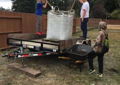 Two students stand on a trailer with a big bag of soil read to shovel soil into planter boxes